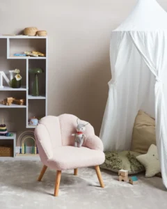 Vaaleanpunainen pehmeä lasten nojatuoli puisilla jaloilla vaaleassa lastenhuoneessa lelujen ja leikkiteltan vieressä.